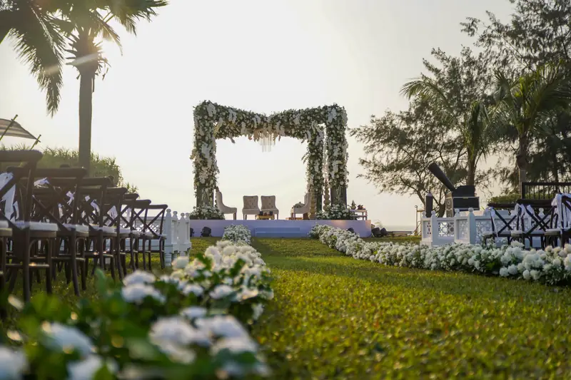 A white floral wedding mandap at the end of a sunlit lawn aisle overlooking the sea